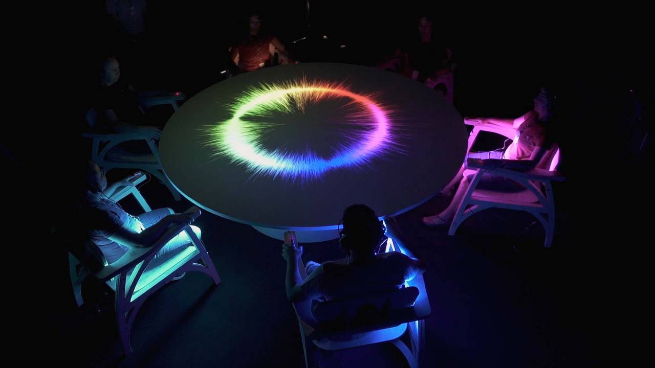 Pessoas sentadas em torno de uma mesa redonda com tudo escuro e um círculo de luzes coloridas em cima da mesa