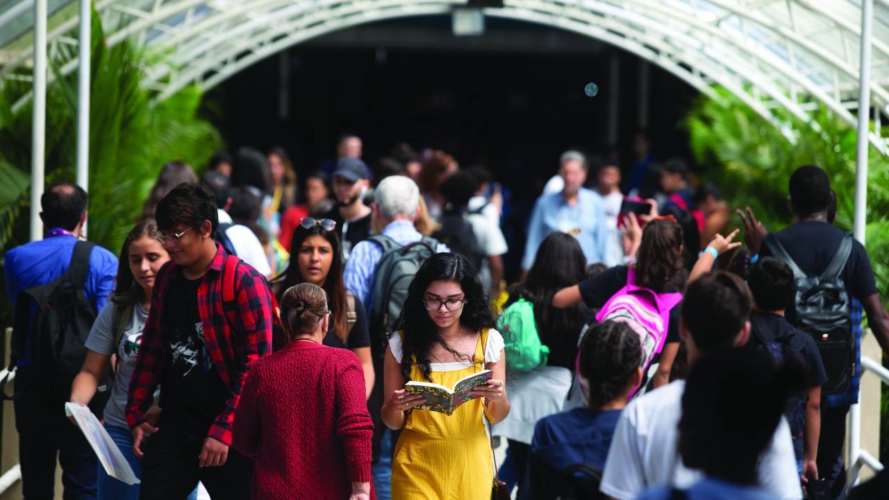 Multidão andando em um corredor da Bienal, com uma jovem de amarelo lendo um livro. Ela é uma mulher branca, de cabelo escuro ondulado e usa óculos