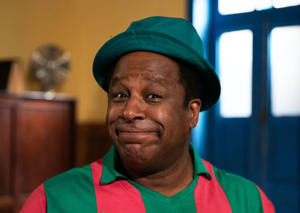 Ailton Graça está caracterizado como Mussum. Ele é um homem negro de pele escura, usa chapéu estilo "bucket hat" azul e blusa de gola polo listrada verde e rosa