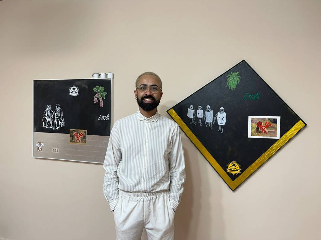 Jaime Lauriano é um homem negro de pele clara. Ele está de camisa branca listrada e calça branca, sorrindo e com as mãos no bolso. Tem o cabelo curto e descolorido, barba, bigode e cavanhaque pretos. Atrás dele, estão dois quadros: um é retangular e o outro é quadrado, com um dos vértices para cima. Os dois com fundo preto.