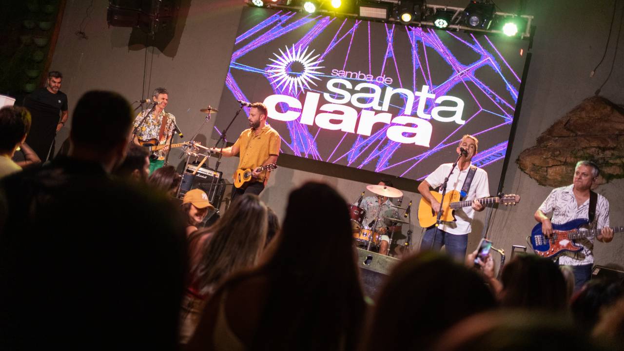 Na frente da foto, as silhuetas de pessoas da plateia. No fundo, o palco, com os seis integrantes do grupo Samba de Santa Clara. O palco tem o nome Santa Clara projetado em letras brancas grandes.