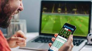 Homem confere celular enquanto TV exibe jogo de futebol.