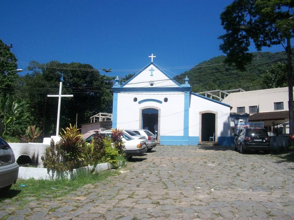 A imagem é uma fotografia de uma capela azul e branca muito simples. Há carros parados em frente.