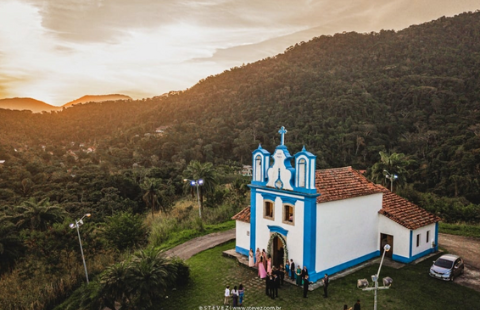 A imagem é uma fotografia aérea que mostra uma bela paisagem, incluindo uma igreja colonial azul e branca, com pessoas em frente a ela, no que parece ser um casamento. Na colina, há vegetação tropical e um belo entardecer.