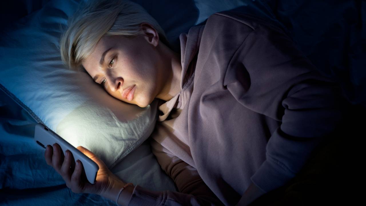 Pessoa deitada na cama mexe no telefone celular.