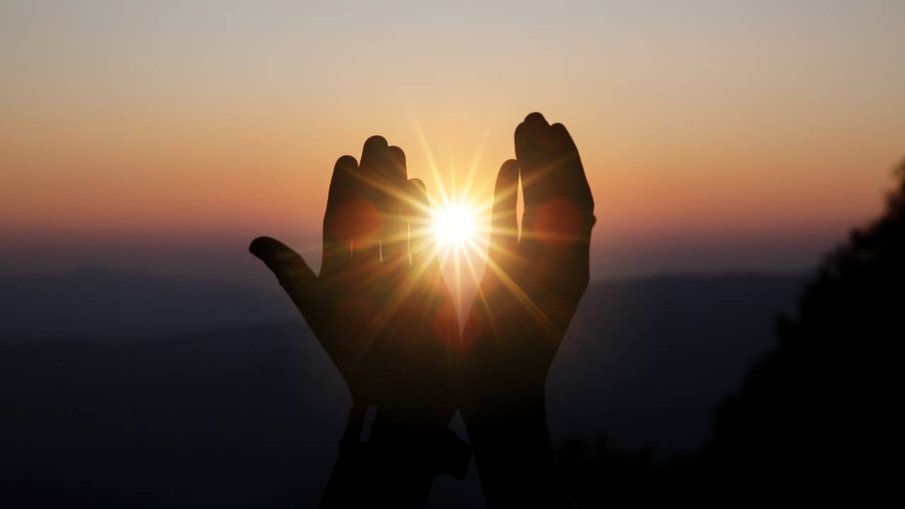 Mãos em postura de reza tendo o sol ao fundo.