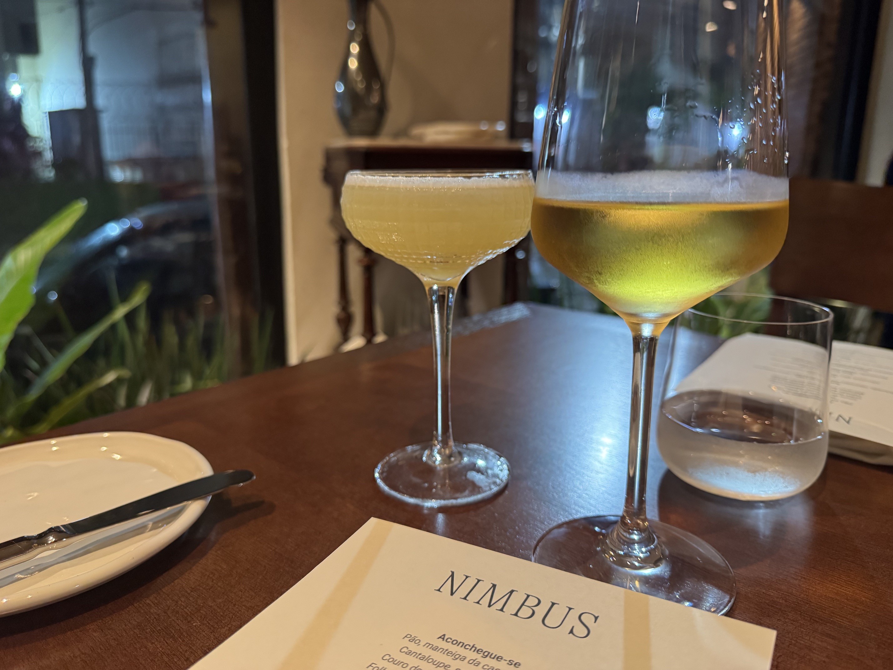 Mesa de madeira com dois copos de bebida amarela, um prato branco com talheres e um cardápio
