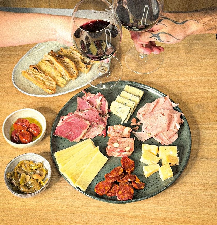 Duas pessoas brindam com taças de vinho tinto sobre uma mesa de madeira, com uma tábua de frios, pães e molhos. Ao fundo, garrafas de vinho com rótulos variados.