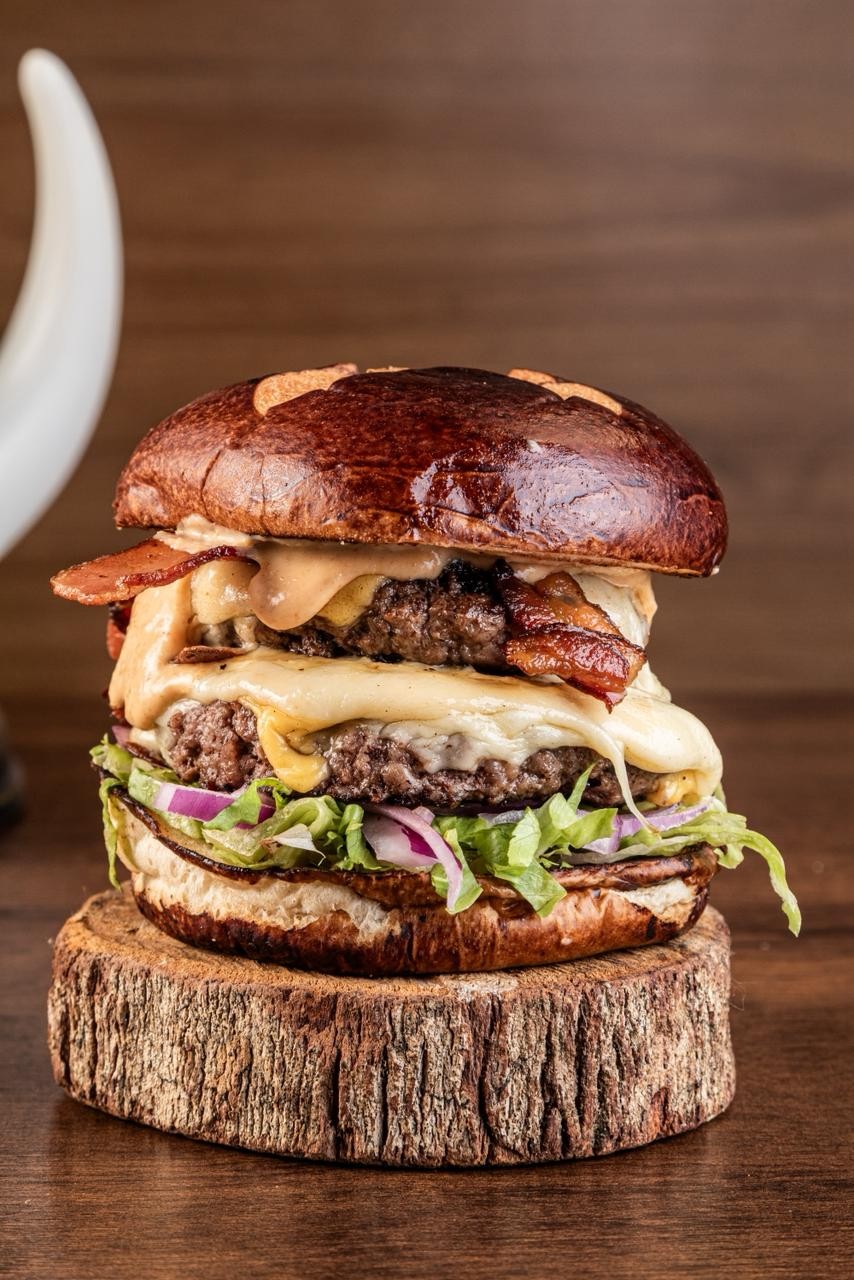 Hambúrguer duplo com pão brioche escuro, dois hambúrgueres de carne, queijo derretido, bacon crocante, alface e cebola roxa, servido sobre um tronco de madeira rústico em uma mesa escura