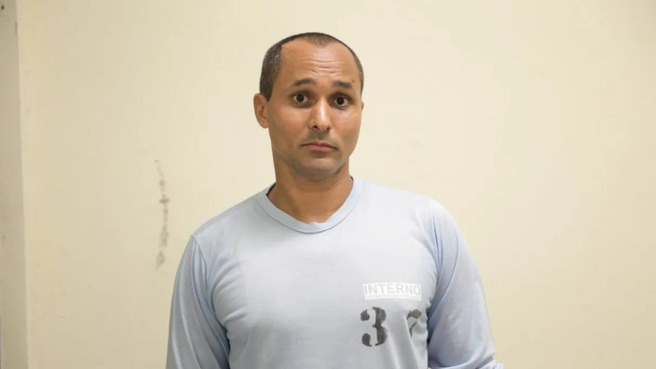 Homem de pele parda, cabelo curto e ralo, olhos arregalados, boca fechada, vestindo camiseta azul clara de manga comprida com a palavra "INTERNO" e o número "32" no peito, em frente a uma parede bege clara