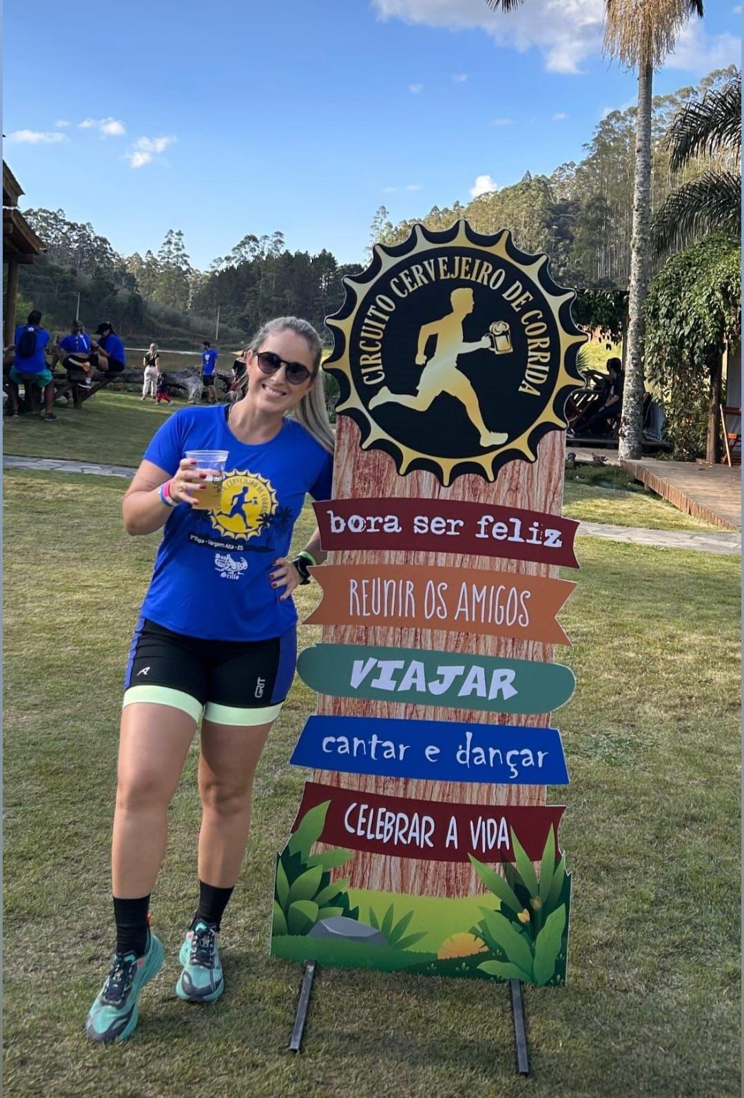 Mulher loira sorridente, de óculos escuros, camiseta azul e shorts de corrida, segura um copo de cerveja e posa ao lado de uma placa de madeira com a frase 