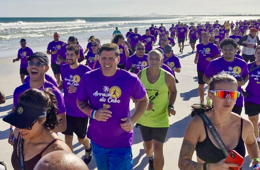 Multidão de pessoas correndo na areia da praia, a maioria vestindo camisetas roxas com a inscrição 