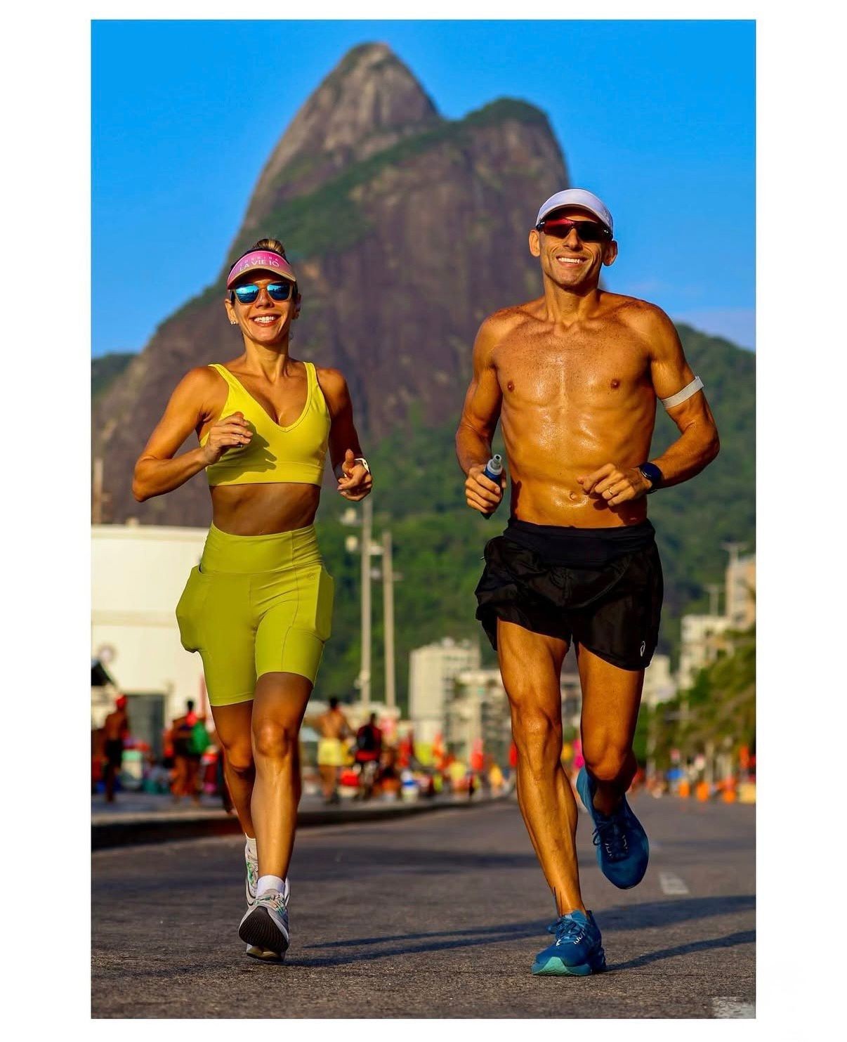 Casal sorridente correndo na rua, mulher de top e bermuda amarela, homem sem camisa de shorts preto, ambos com óculos escuros e bonés. Ao fundo, montanha rochosa e prédios sob céu azul