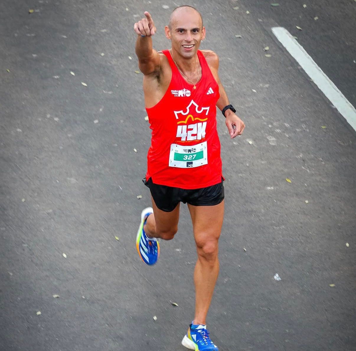 Corredor careca, sorrindo, vestindo regata vermelha com número 327 e shorts pretos, apontando para cima enquanto corre em uma rua cinza