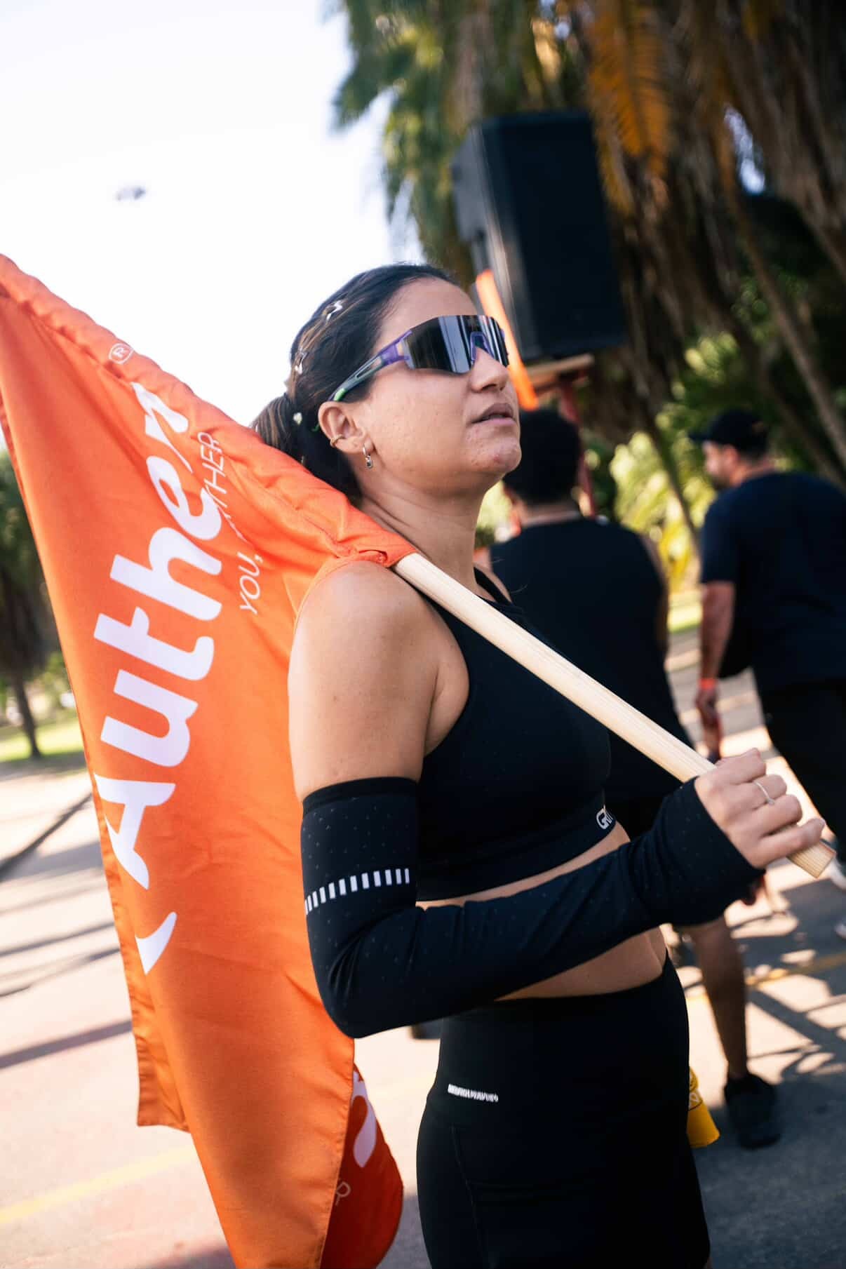 Mulher de óculos escuros e roupa esportiva preta, com uma braçadeira preta e branca no braço esquerdo, segura uma bandeira laranja com a palavra 