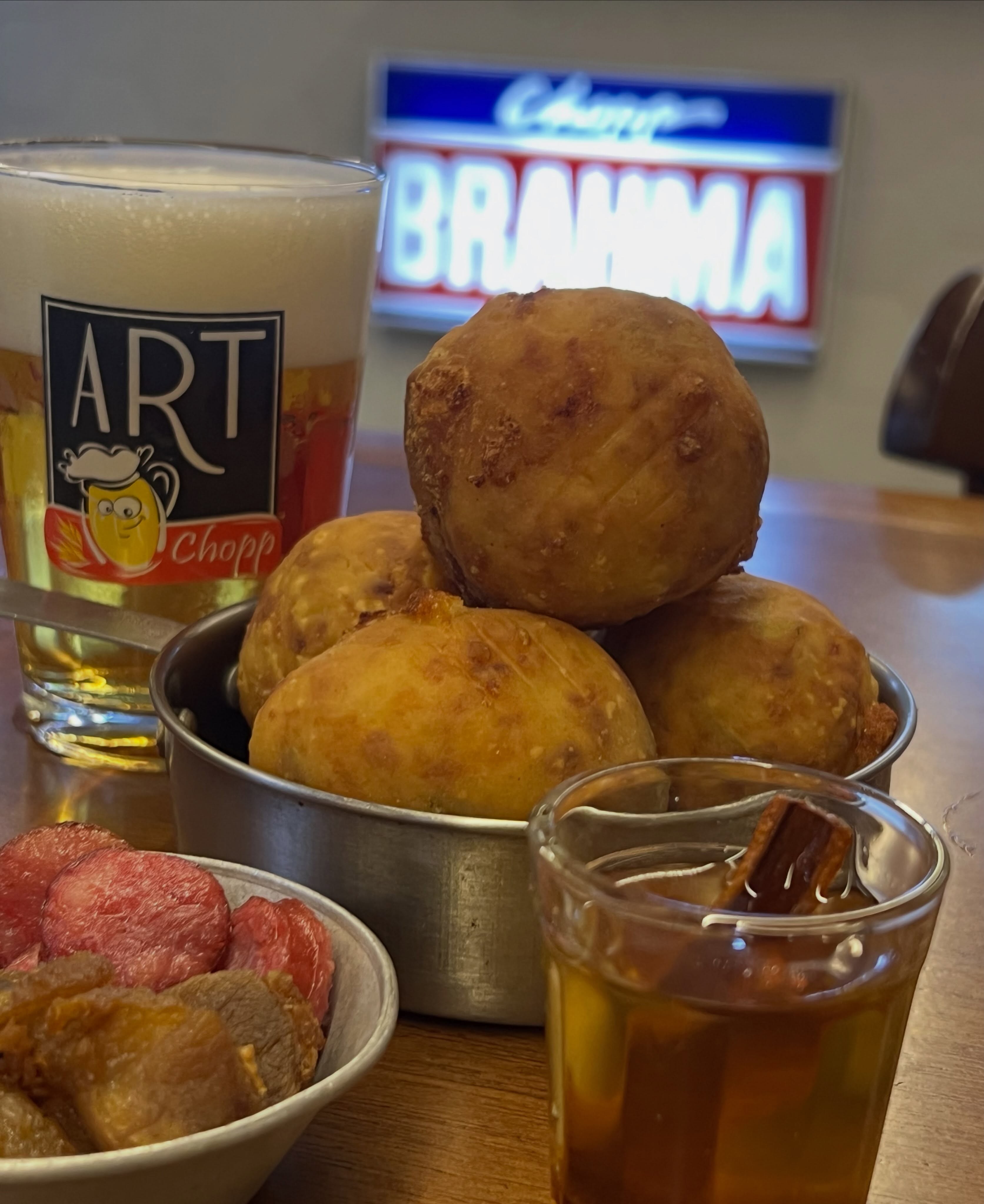 Copo de chopp Art Chopp com espuma, tigela de bolinhos fritos, por&ccedil;&atilde;o de lingui&ccedil;a e carne, e um shot de cacha&ccedil;a com canela, sobre mesa de madeira, com letreiro neon Brahma ao fundo