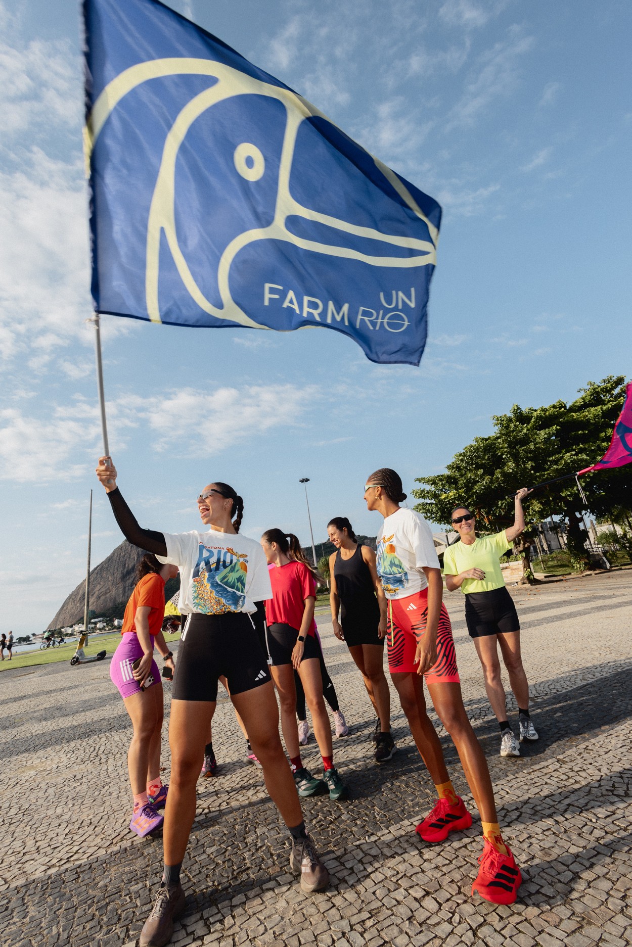 Mulher sorridente de óculos escuros e camiseta branca com estampa colorida, segurando uma bandeira azul com logo Farm Rio, em meio a um grupo de mulheres em roupas esportivas, em calçadão de paralelepípedos com céu azul e montanhas ao fundo