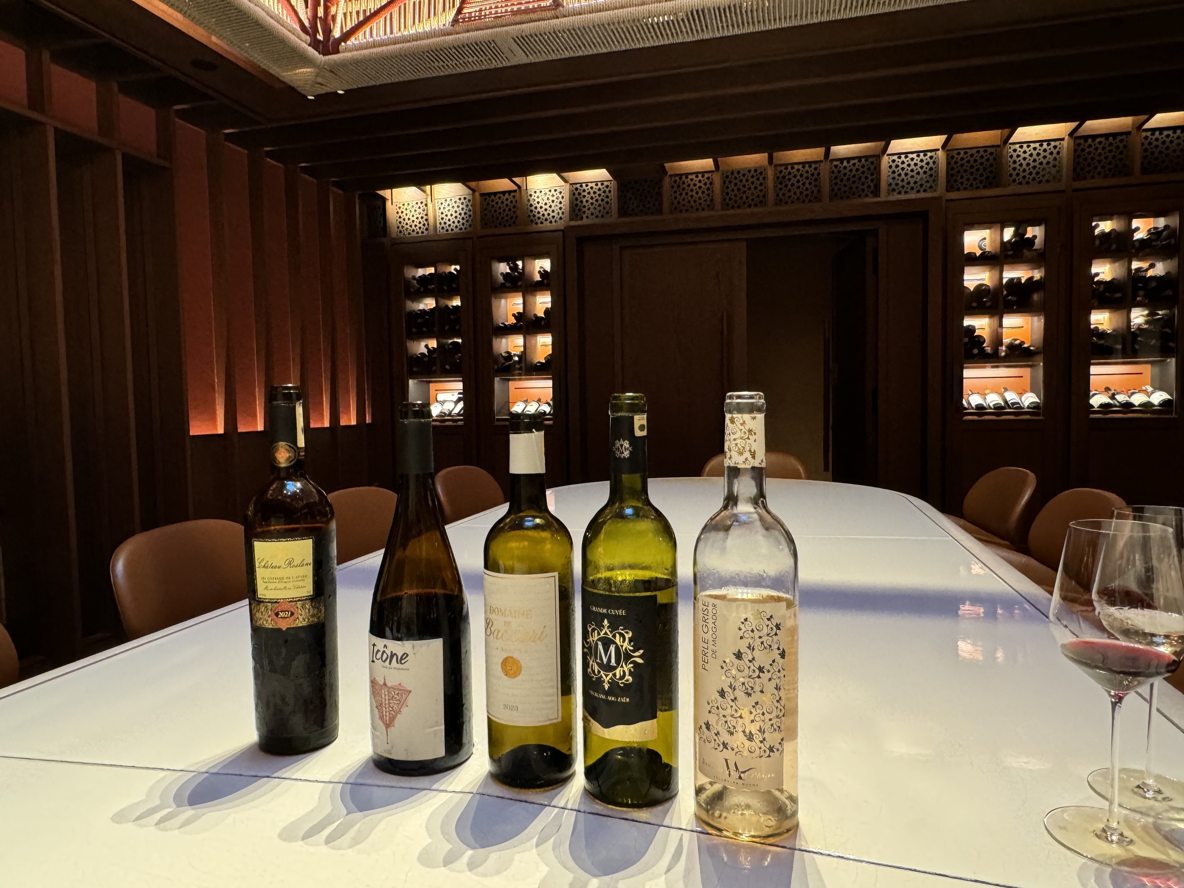 Cinco garrafas de vinho, três brancas e duas tintas, alinhadas sobre uma mesa branca, com duas taças de vinho tinto ao lado direito. Ao fundo, uma adega com prateleiras de madeira escura e garrafas de vinho iluminadas, criando um ambiente sofisticado