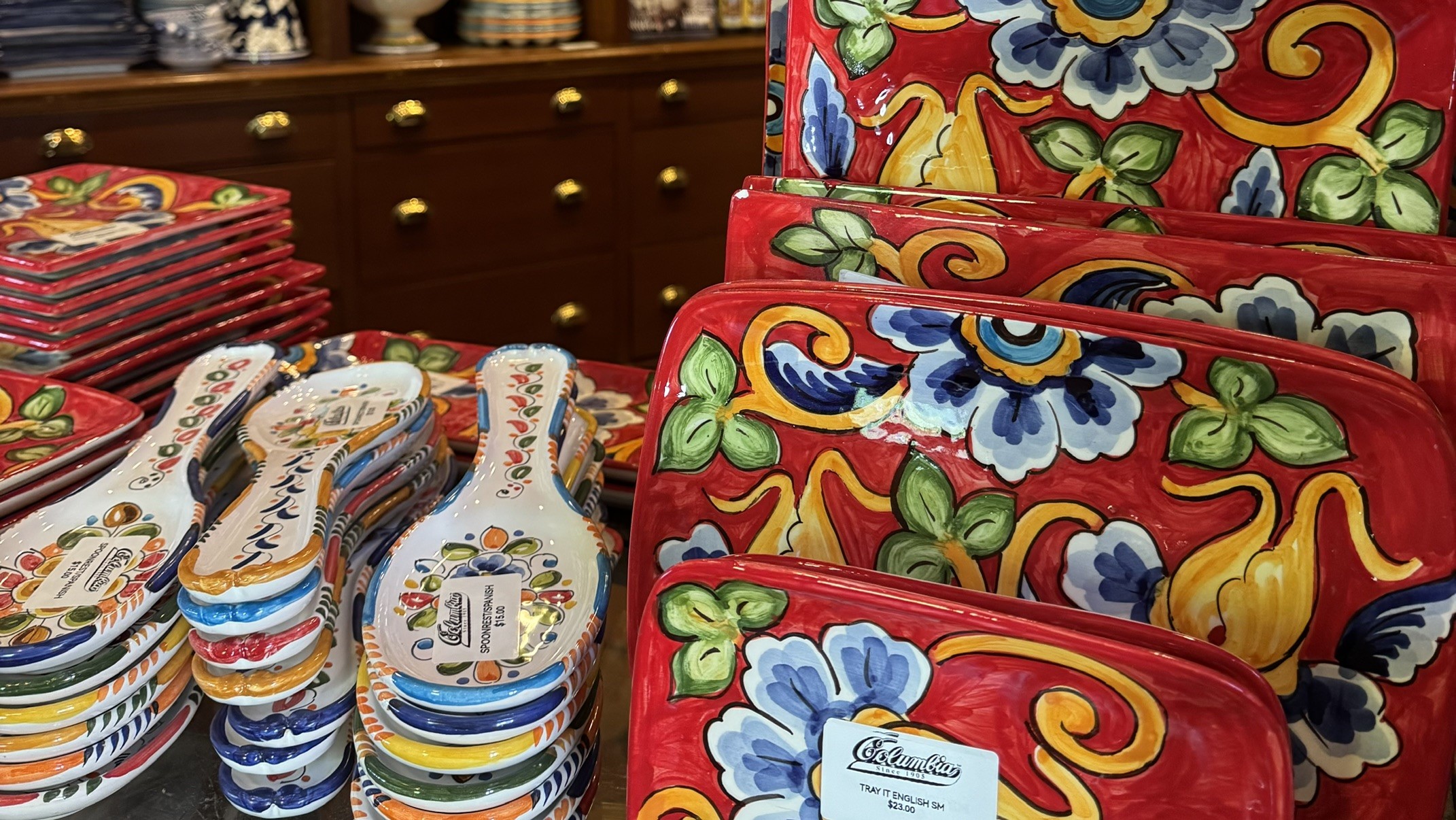 Exposição de cerâmicas artesanais com pratos e apoios de colher vermelhos e brancos, decorados com flores azuis, folhas verdes e arabescos amarelos. Ao fundo, um móvel de madeira escura com gavetas e puxadores dourados, exibindo mais peças de cerâmica