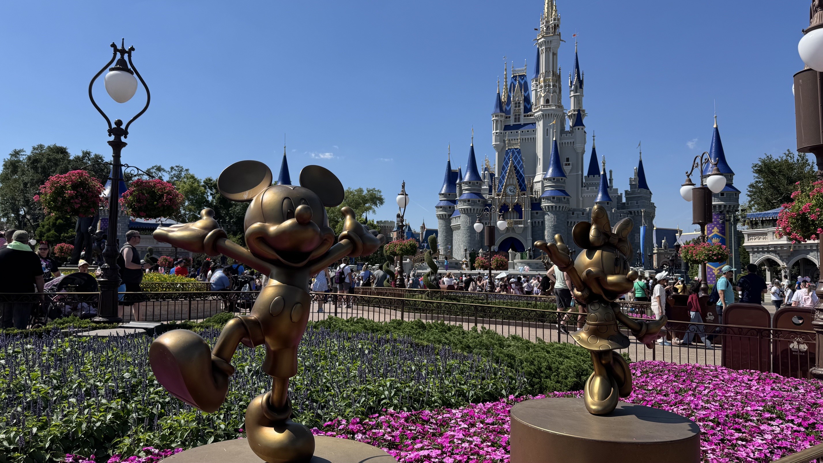 Estátuas douradas de Mickey e Minnie Mouse em um jardim florido, com o Castelo da Cinderela ao fundo e pessoas caminhando em um dia ensolarado
