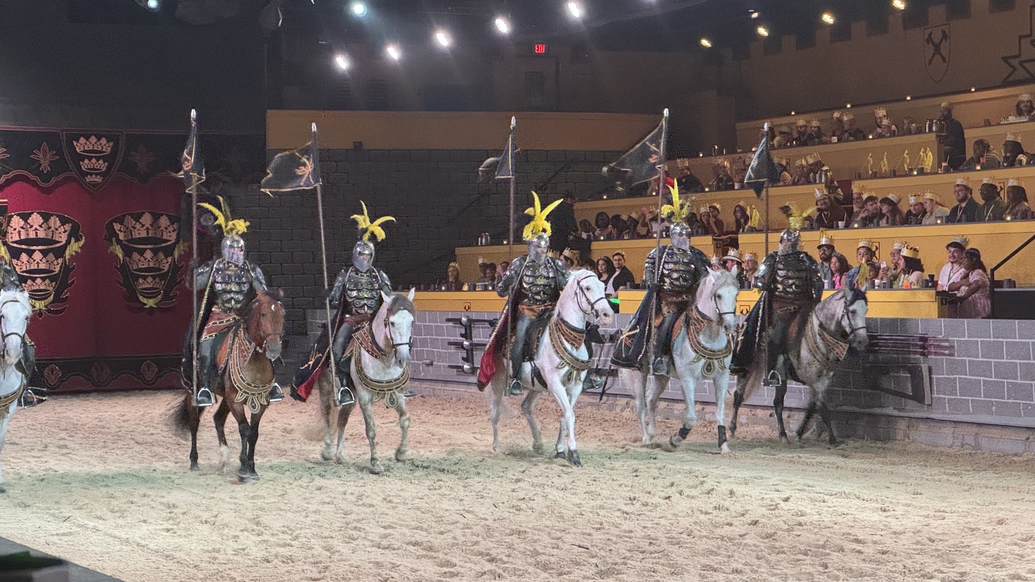 Cavaleiros medievais com armaduras e capacetes emplumados montam cavalos em uma arena de areia, com público em arquibancadas ao fundo