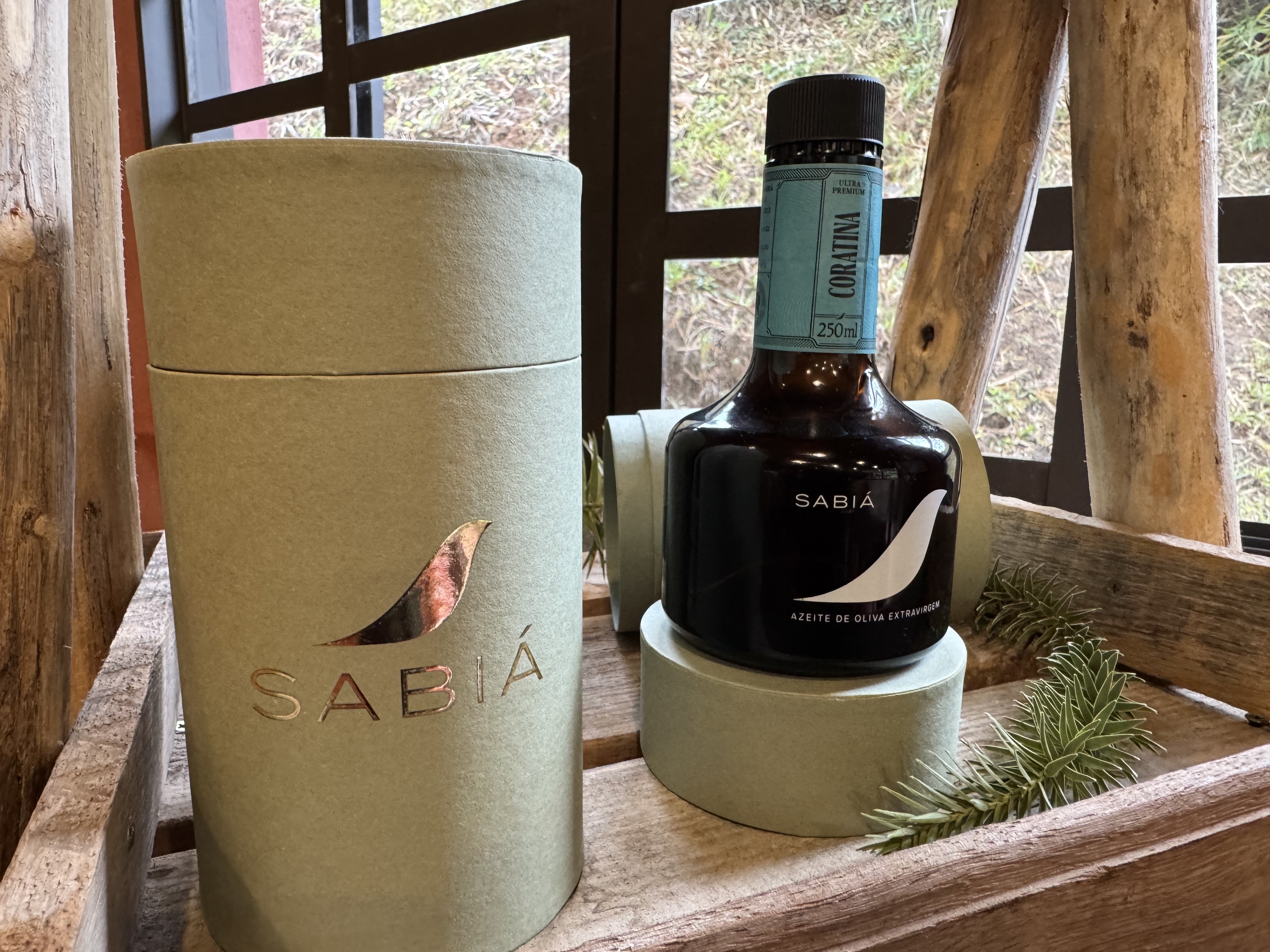 Em primeiro plano, uma caixa cilíndrica verde-clara com o logo Sabiá em dourado e uma garrafa escura de azeite Sabiá Coratina 250ml sobre um suporte redondo, com um ramo verde ao lado. Ao fundo, uma janela com vista para a vegetação