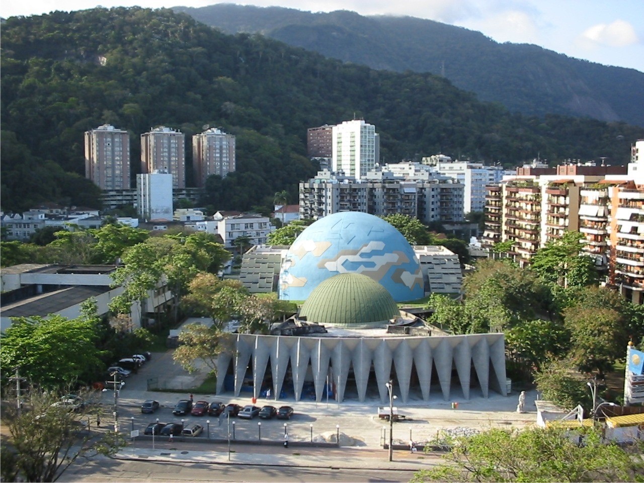 Vista aérea do Planetário do Rio de Janeiro, com sua cúpula azul e verde, cercado por prédios residenciais e árvores. Ao fundo, montanhas verdes sob céu parcialmente nublado