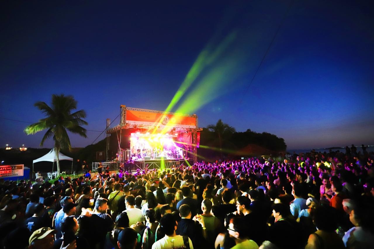 Multidão assiste a um show noturno ao ar livre, com palco iluminado por luzes coloridas e raios de luz verde projetados no céu escuro. Uma palmeira e uma tenda branca aparecem à esquerda