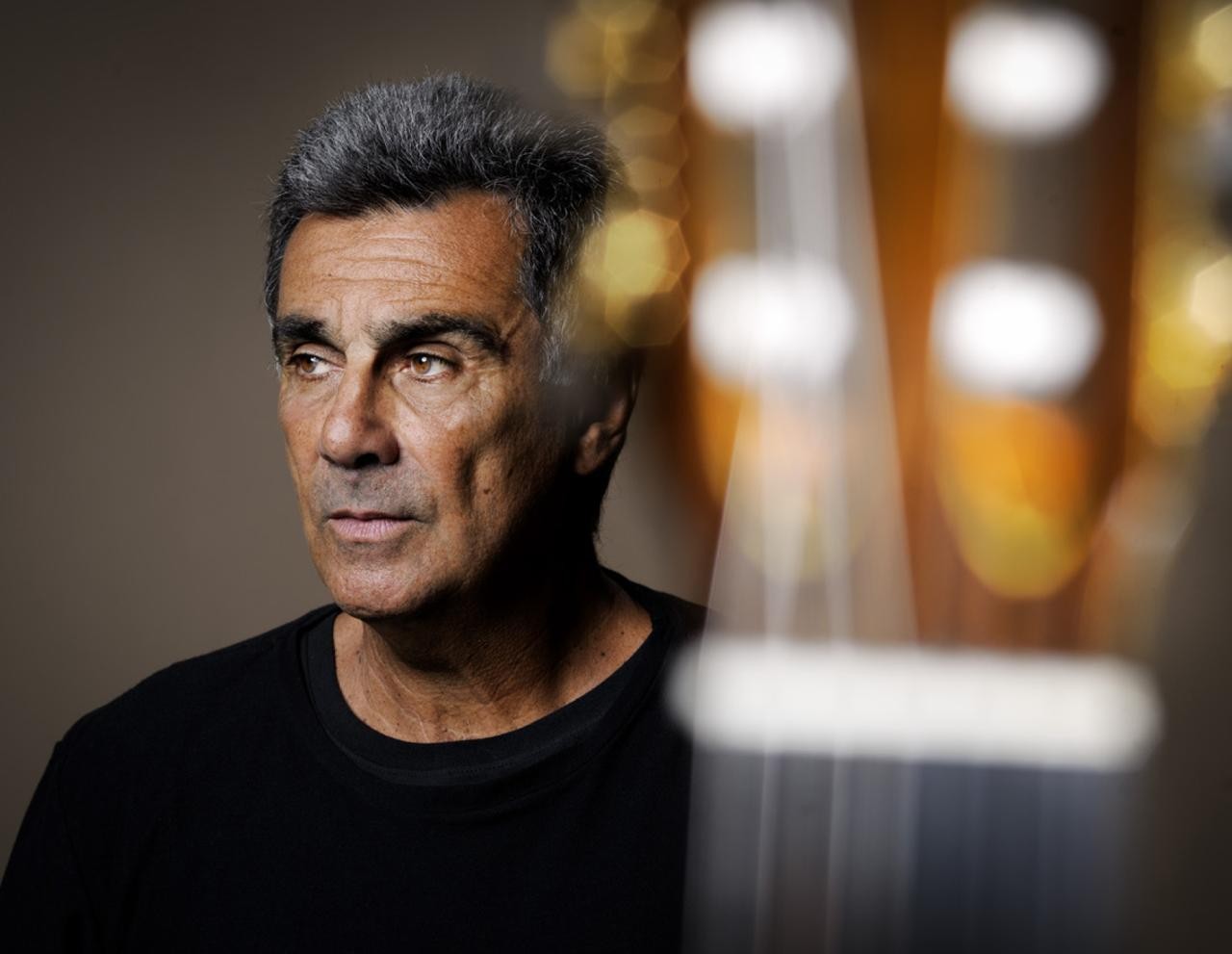 Homem de pele bronzeada, cabelo grisalho e camiseta preta, olhando pensativo para a esquerda, com um instrumento musical desfocado à direita