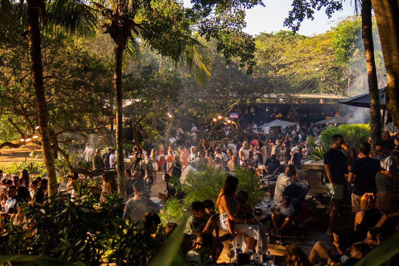 Multidão de pessoas em um evento ao ar livre, com palmeiras e vegetação densa. Há fumaça no ar, luzes penduradas e barracas, sugerindo uma festa ou festival em meio à natureza