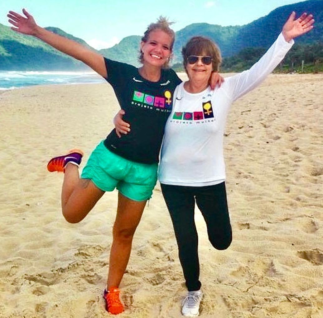 Duas mulheres sorridentes, uma jovem e uma idosa, abraçadas na praia. A jovem veste camiseta preta e shorts verde, levantando uma perna. A idosa, de óculos escuros, usa camiseta branca de manga longa e calça preta. Ambas exibem camisetas com a estampa projeto mulher e símbolos femininos coloridos. Ao fundo, o mar e montanhas verdes sob um céu azul