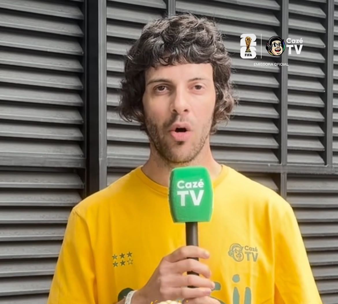 Homem de cabelo e barba castanhos, vestindo camiseta amarela, segura um microfone verde da Caz&eacute; TV e tem a boca aberta, como se estivesse falando. Ao fundo, uma parede cinza com persianas horizontais. No canto superior direito, o logo da FIFA e da Caz&eacute; TV, com a inscri&ccedil;&atilde;o Emissora Oficial. Na camiseta, tr&ecirc;s estrelas douradas e o logo da Caz&eacute; TV.