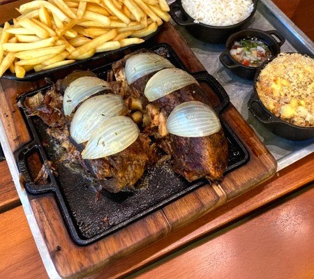Prato de carne assada com rodelas de cebola em chapa de ferro, acompanhado de batatas fritas, arroz branco, vinagrete e farofa em tigelas pretas, sobre uma bandeja met&aacute;lica e t&aacute;bua de madeira