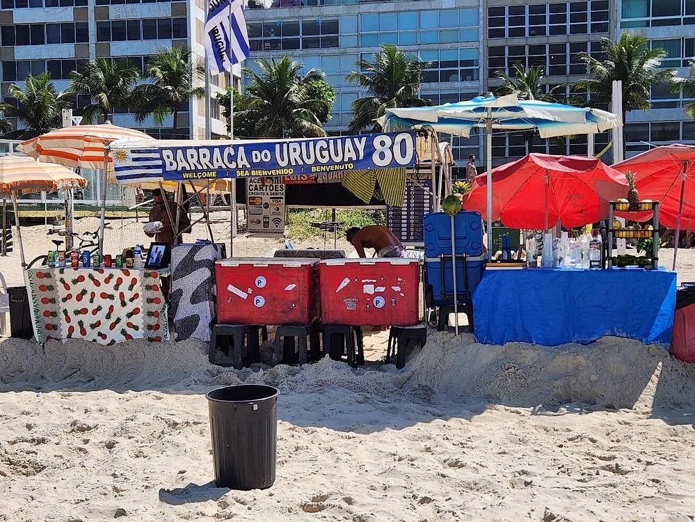 Barraca do Uruguai 80 na praia, com bandeira uruguaia, guarda-s&oacute;is coloridos, mesas com bebidas e frutas, e um homem trabalhando. Ao fundo, pr&eacute;dios e palmeiras sob c&eacute;u azul