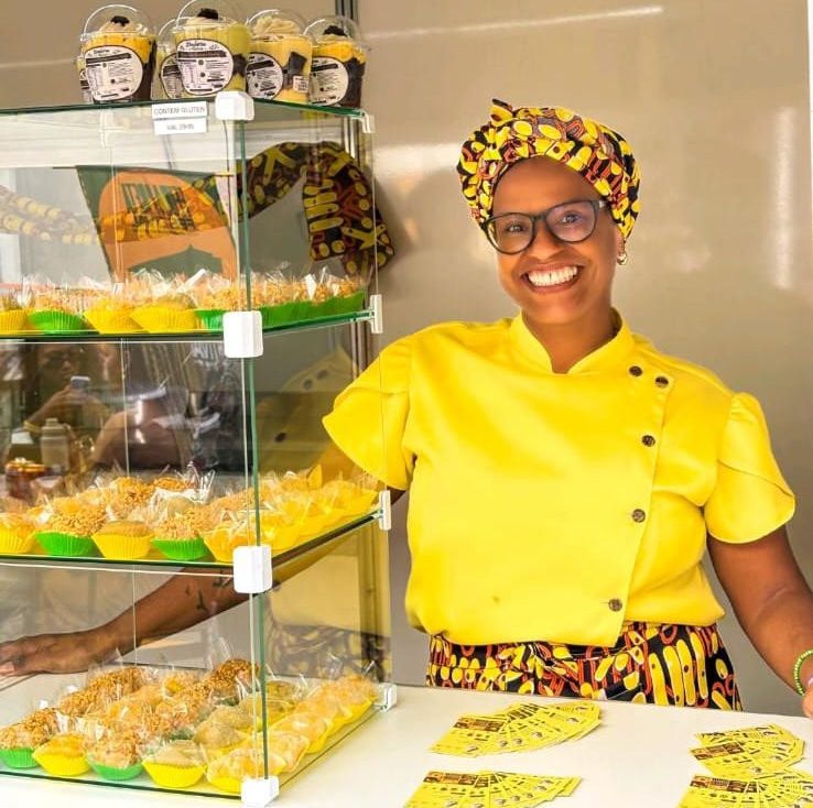 Mulher negra sorridente, de óculos e turbante estampado amarelo, veste dólmã amarela e saia estampada, atrás de um balcão com doces em potes e forminhas, e cartões de visita amarelos