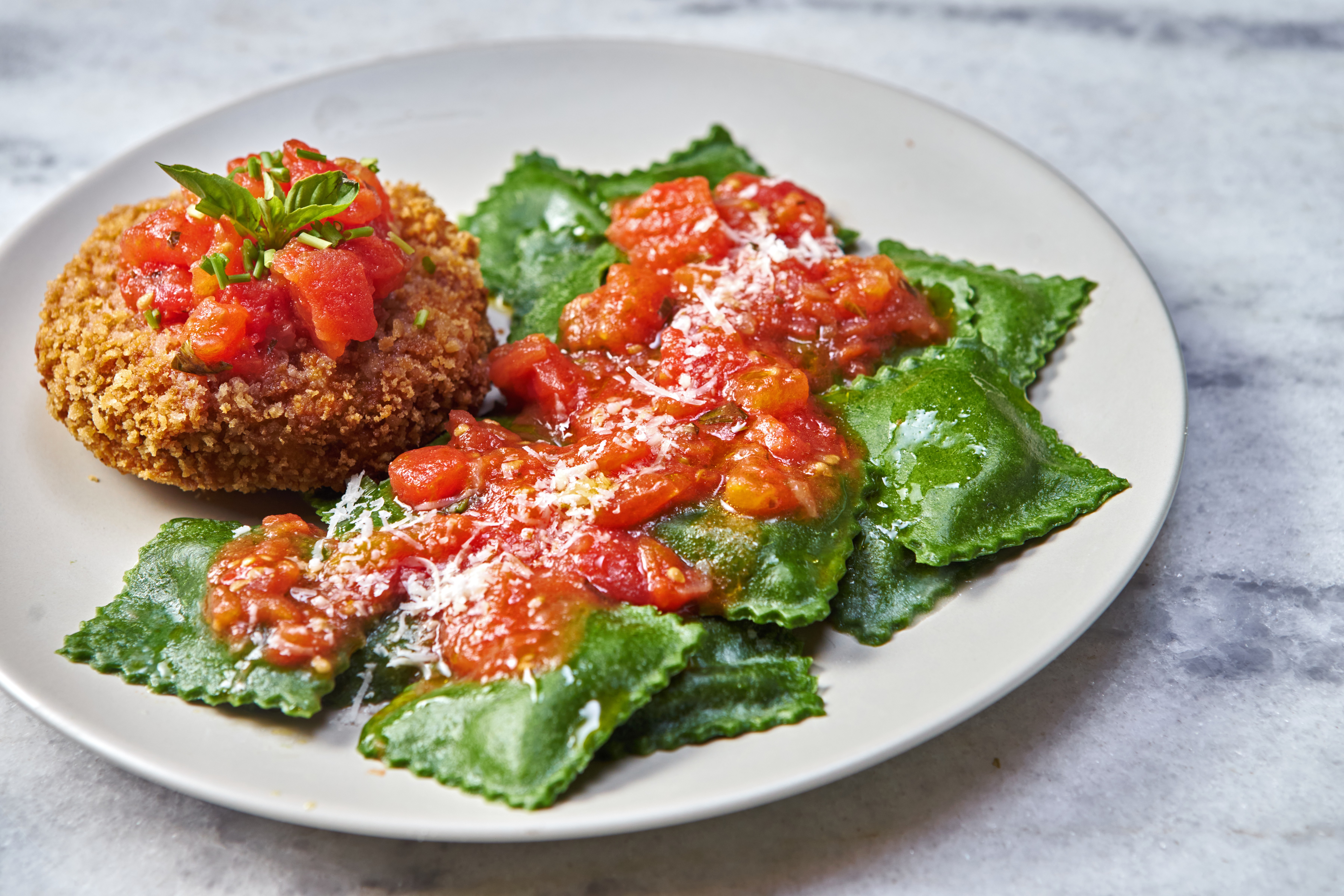 Prato com um bolinho frito dourado coberto por molho de tomate e manjericão, ao lado de raviólis verdes com molho de tomate e queijo ralado