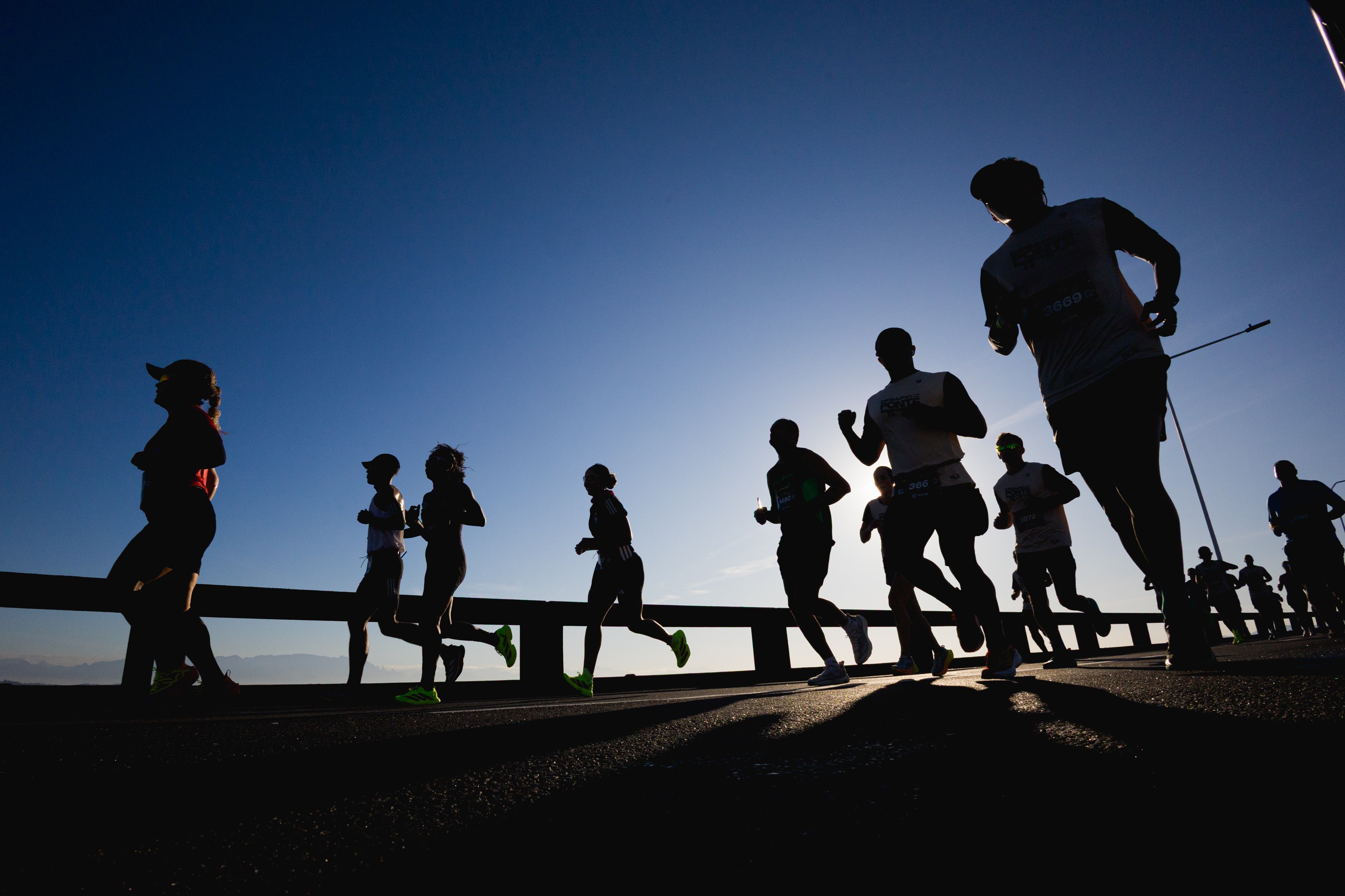 Silhuetas de corredores em uma maratona ao amanhecer, com o sol baixo no horizonte e o céu azul claro. Vários atletas correm em fila, alguns com bonés, em uma ponte ou estrada elevada.