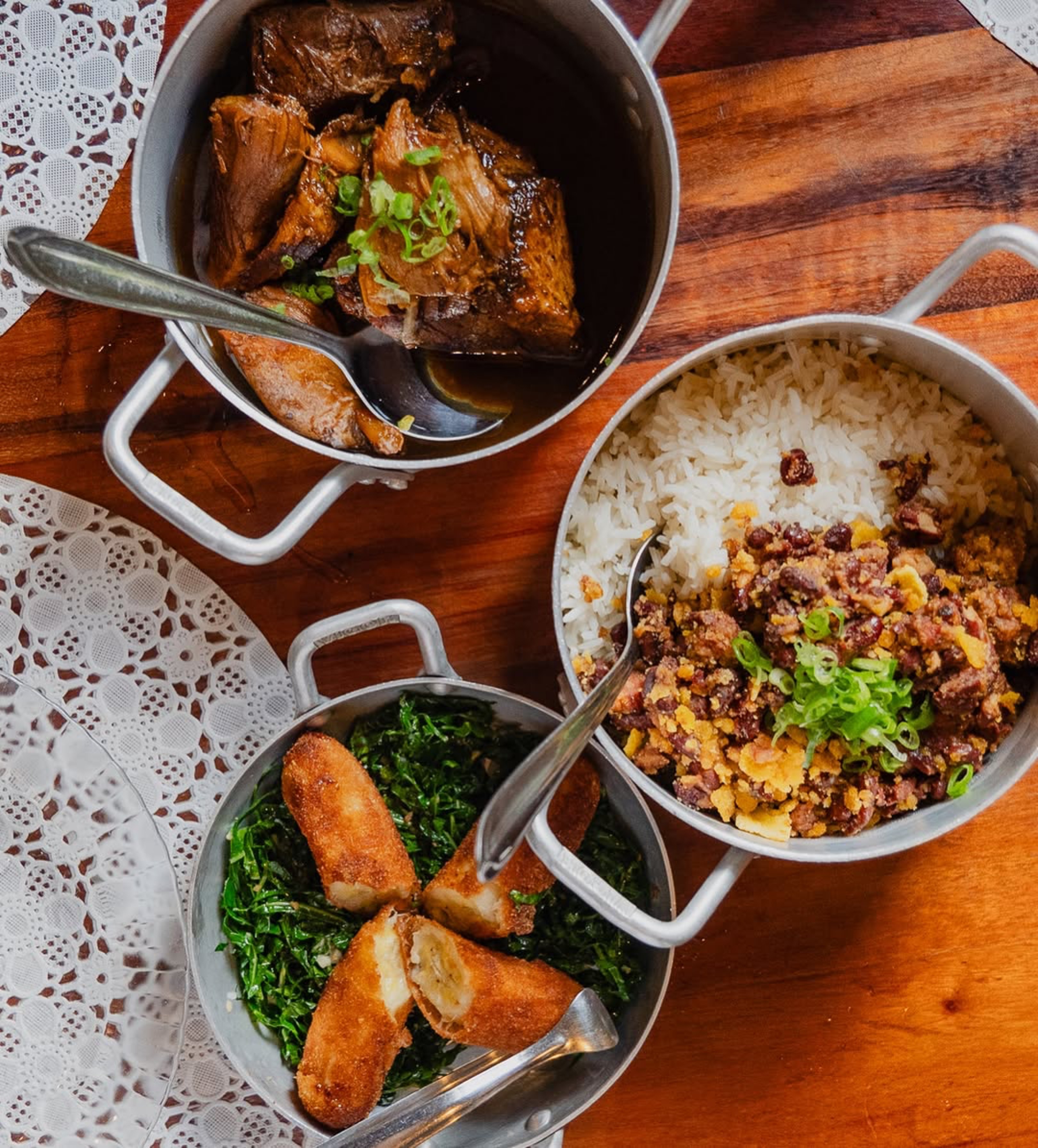 Três panelas de alumínio com comida brasileira sobre uma mesa de madeira. A panela superior tem carne de panela com cebolinha, a do meio tem arroz branco com feijão tropeiro e a inferior tem couve refogada com banana à milanesa