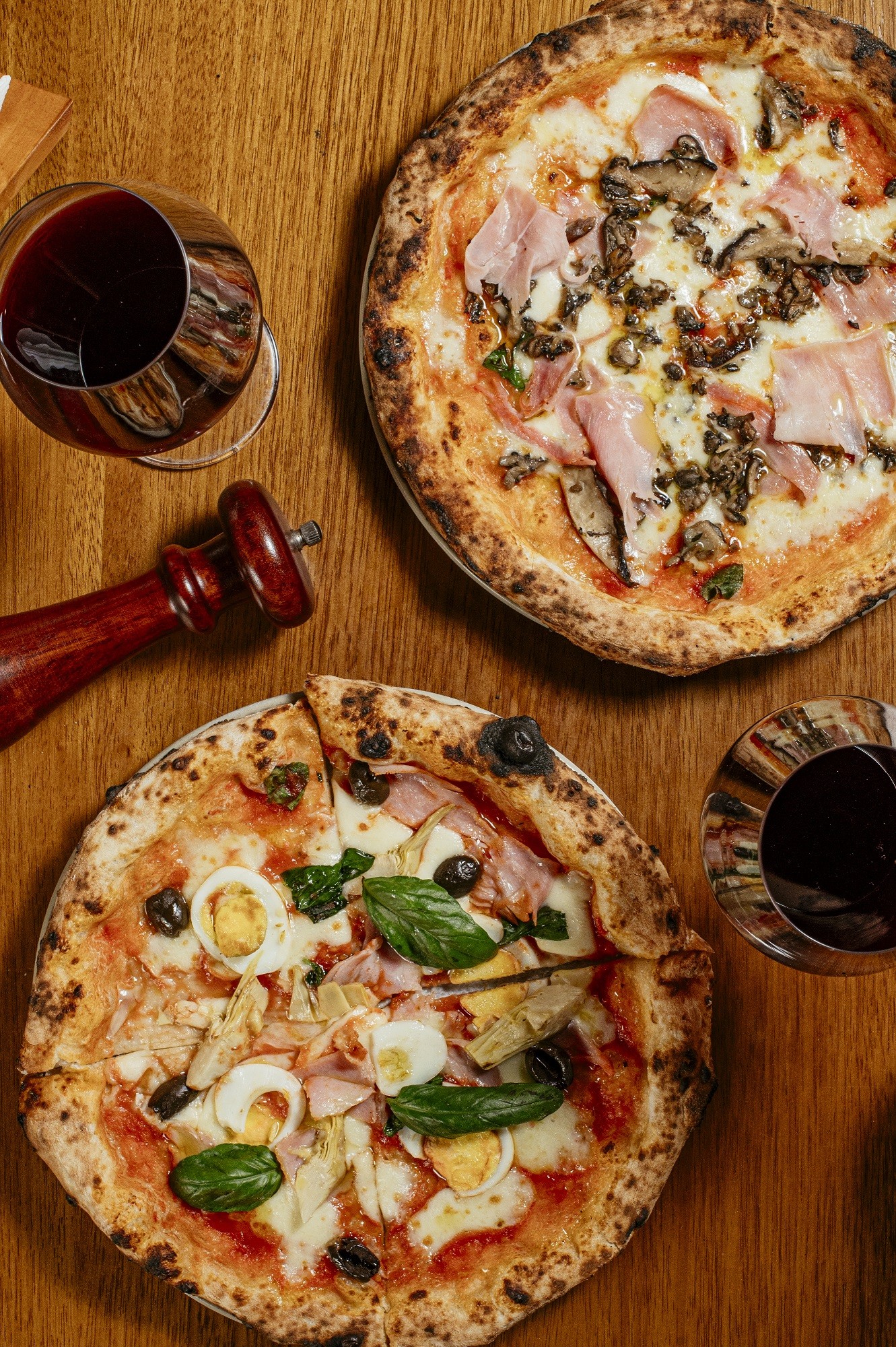 Duas pizzas redondas com bordas crocantes em uma mesa de madeira. A superior tem presunto, cogumelos e manjericão. A inferior, fatiada, tem presunto, ovos de codorna, azeitonas pretas, alcachofras e manjericão. Dois copos de vinho tinto e um moedor de pimenta vermelho completam a cena.