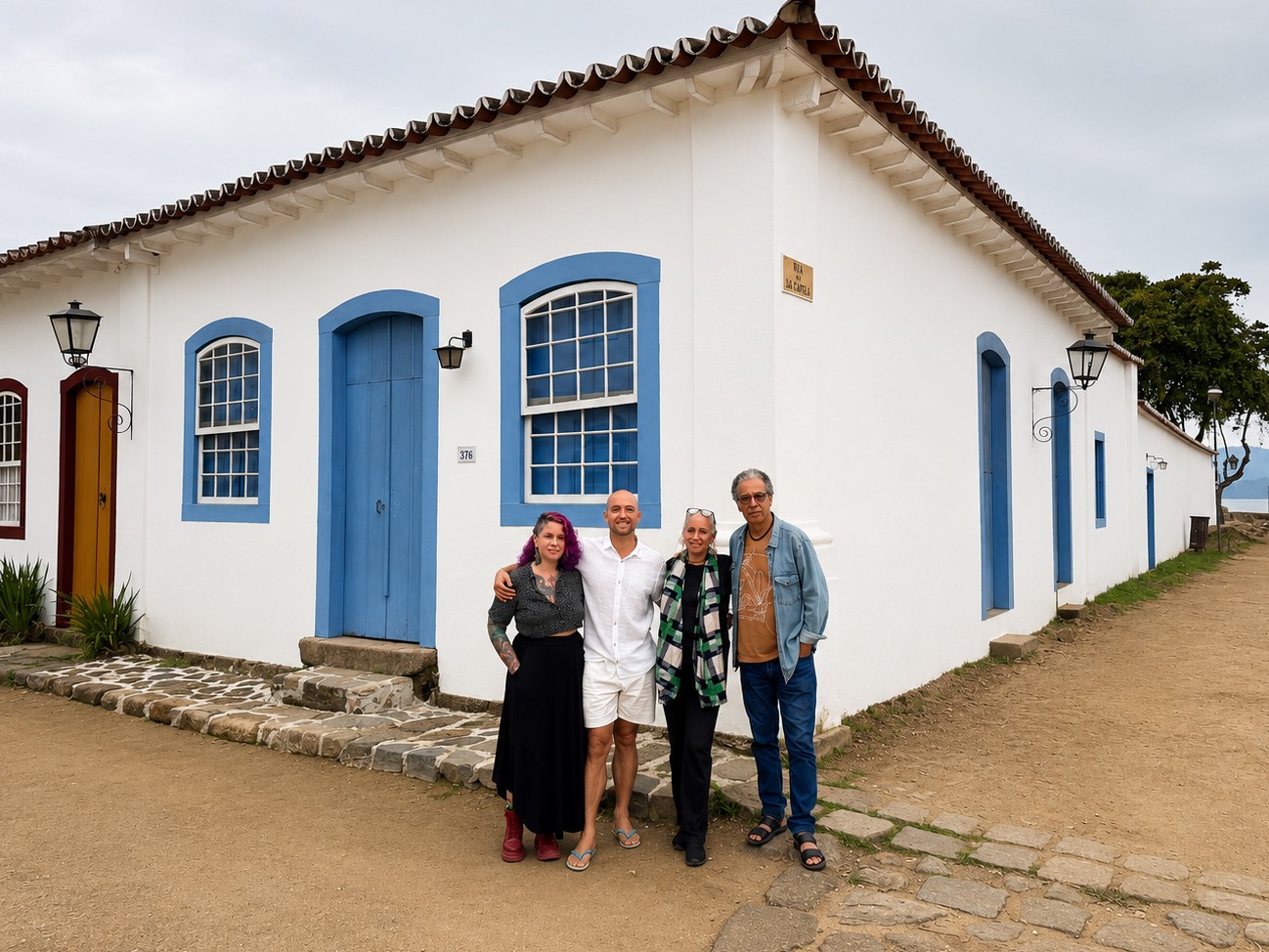Quatro pessoas sorridentes posam em frente a uma casa colonial branca com portas e janelas azuis em Paraty. Da esquerda para a direita: uma mulher de cabelo roxo e saia preta, um homem de camisa branca e bermuda, uma mulher de cabelo grisalho e cachecol xadrez, e um homem de óculos e jaqueta jeans. O chão é de terra e pedras, com o mar visível ao fundo