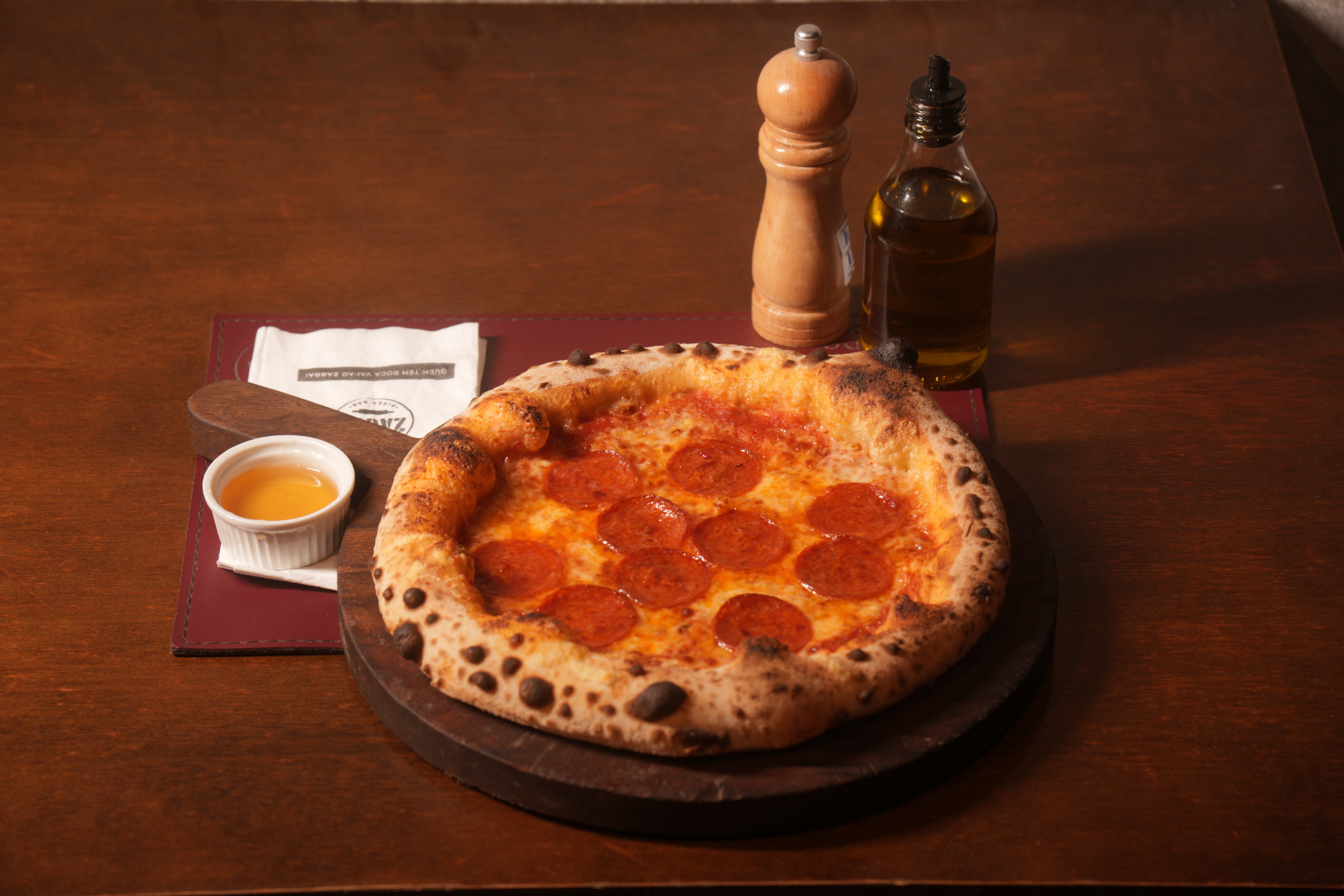 Pizza de pepperoni com borda alta e crocante, servida em tábua de madeira escura. Ao lado, um pote branco com molho, um saleiro de madeira e uma garrafa de azeite, sobre um jogo americano vinho em uma mesa de madeira