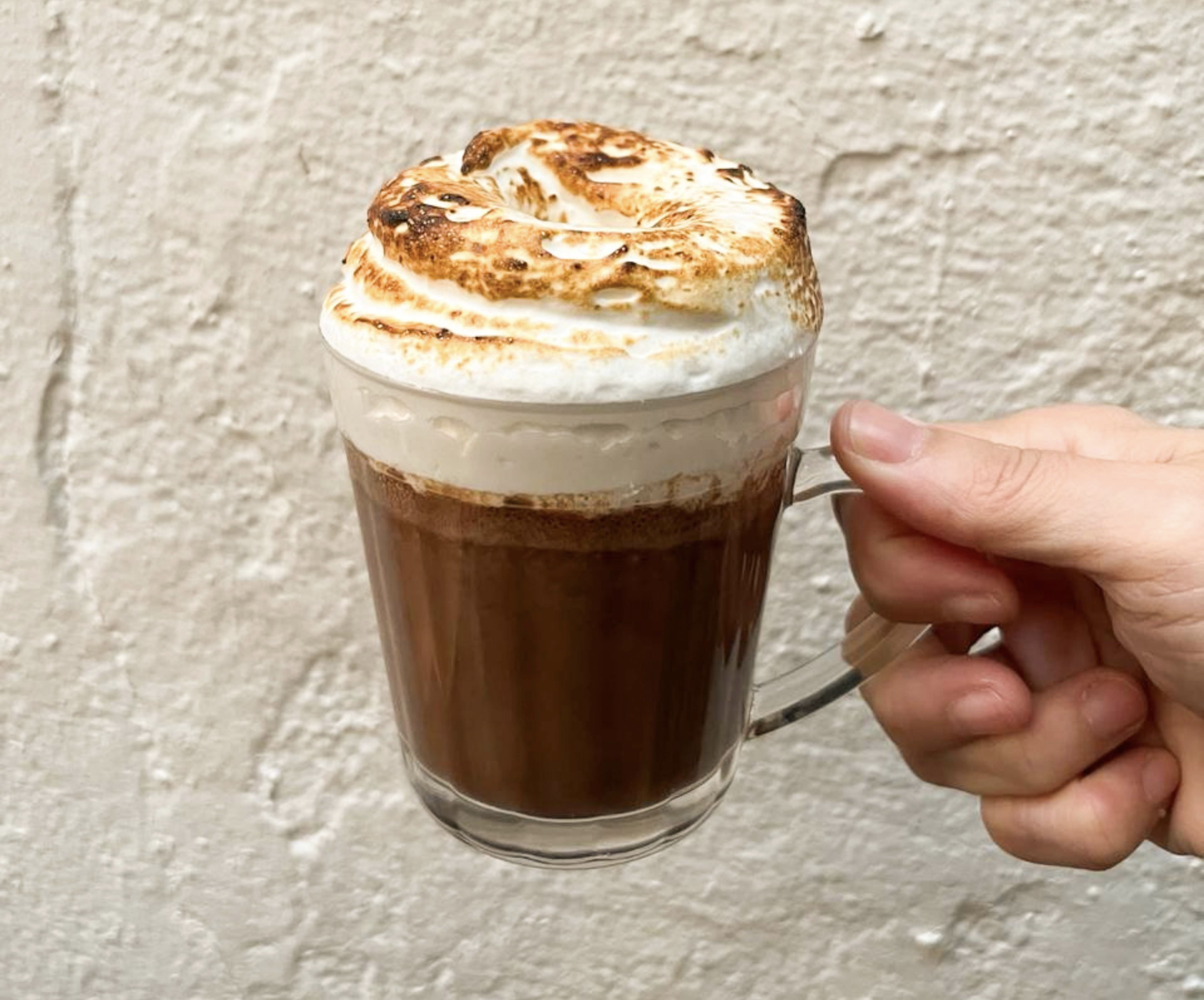 Mão direita segura uma caneca de vidro transparente com chocolate quente e marshmallow tostado por cima, contra uma parede clara texturizada