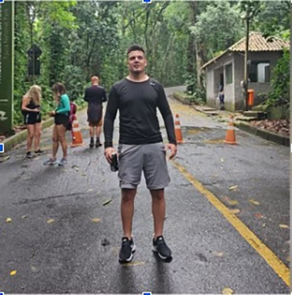 Homem musculoso de camiseta preta e shorts cinza, segurando uma garrafa, em p&eacute; numa estrada molhada com faixa amarela. Ao fundo, outras pessoas e uma guarita em &aacute;rea arborizada