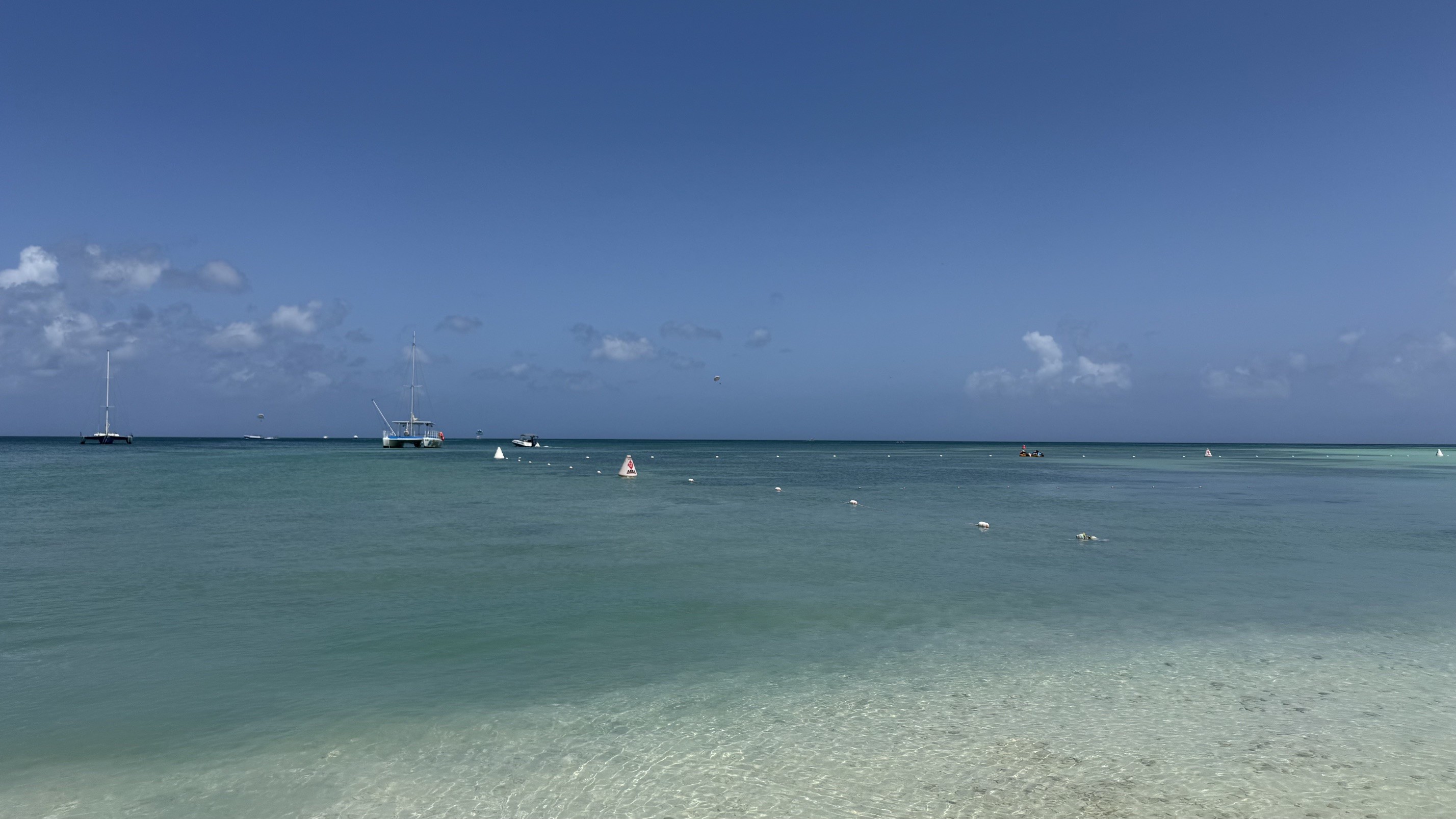 Céu azul com nuvens esparsas sobre um mar de águas cristalinas em tons de azul e verde, com barcos e boias flutuando na distância