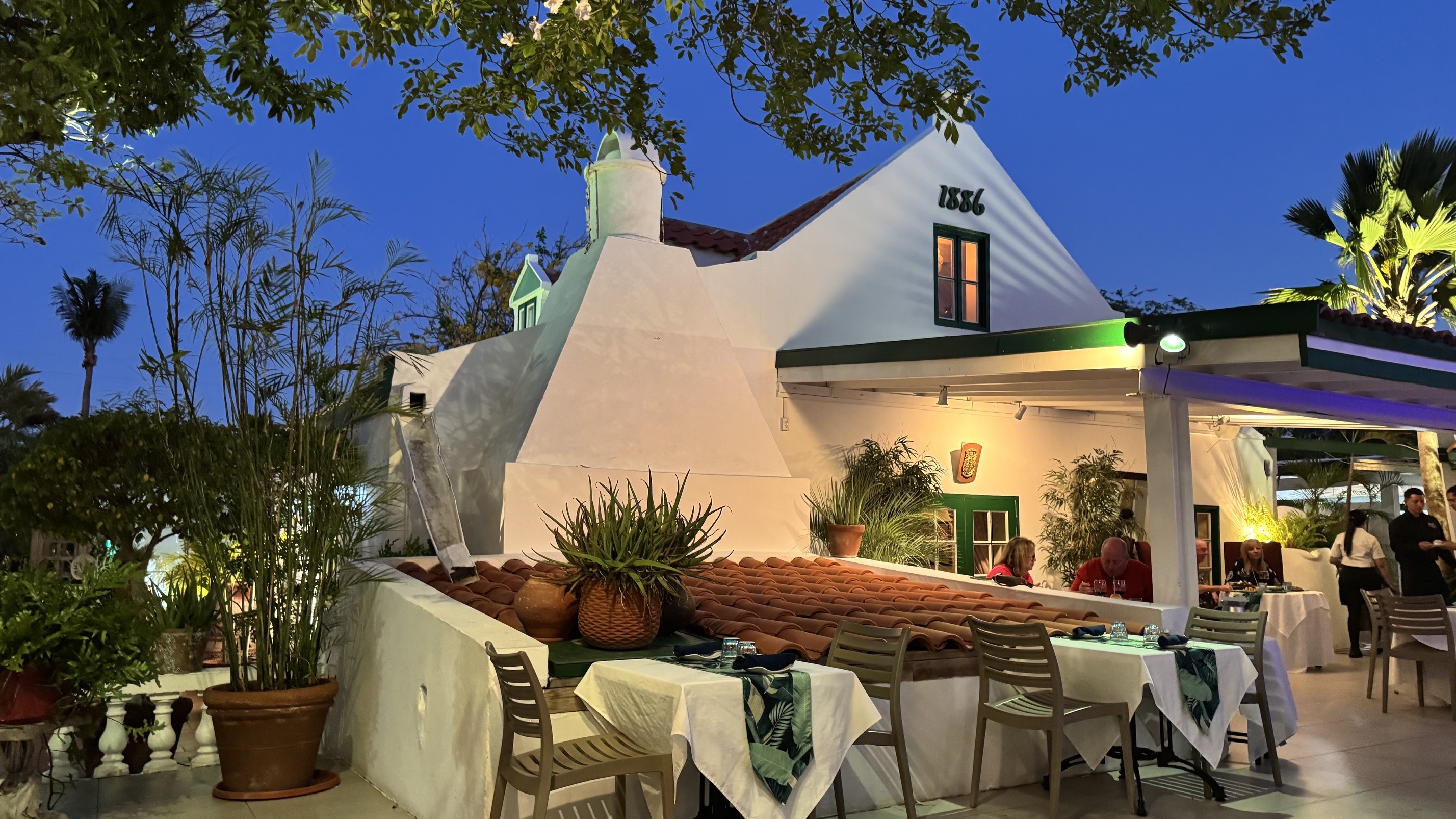 Fachada de um restaurante branco com telhado de terracota e detalhes verdes, iluminado à noite, com mesas postas ao ar livre e pessoas jantando. O céu está azul escuro e há árvores e plantas ao redor.
