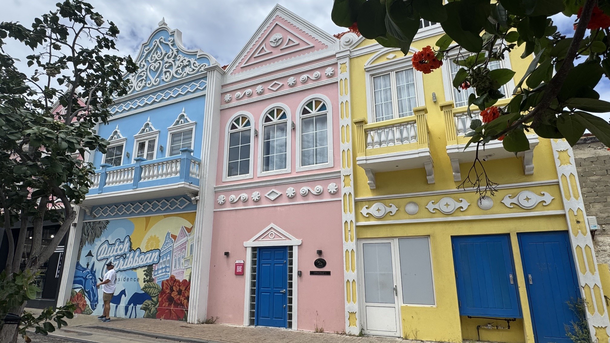 Três edifícios coloridos em estilo colonial holandês. Da esquerda para a direita, um prédio azul, um rosa e um amarelo, todos com janelas brancas e detalhes ornamentados. O prédio azul tem um mural na parte inferior com a frase Dutch Caribbean e um homem de camisa branca e bermuda clara. O prédio rosa tem uma porta azul. O prédio amarelo tem duas portas azuis. Folhagens verdes com flores vermelhas em primeiro plano à direita e à esquerda