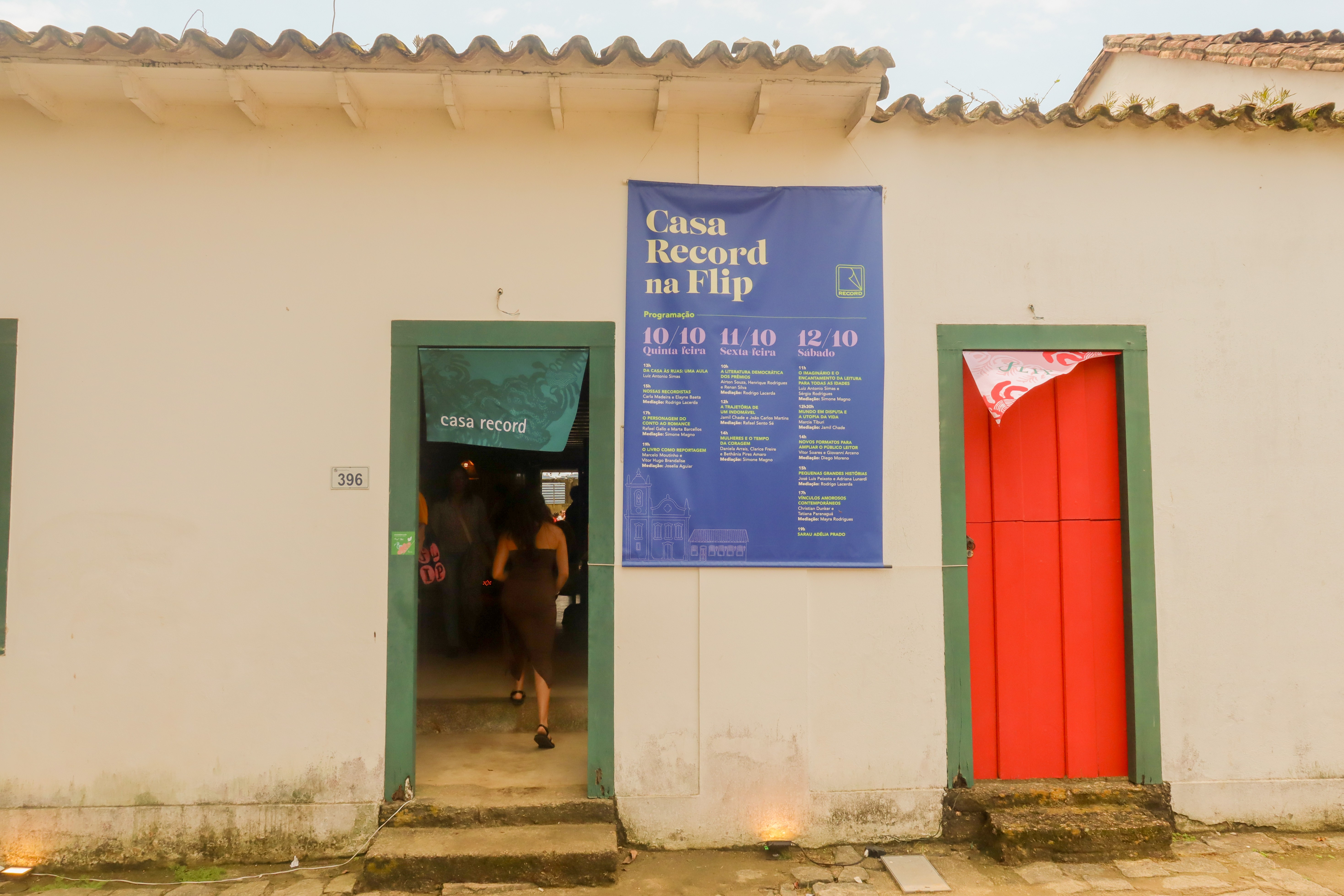Fachada de casa branca com telhado colonial, duas portas com batentes verdes. A porta esquerda est&aacute; aberta, mostrando uma mulher de vestido escuro entrando e uma faixa verde com casa record. Na parede, um banner azul anuncia Casa Record na Flip com programa&ccedil;&atilde;o de 10 a 12 de outubro. A porta direita &eacute; vermelha e est&aacute; fechada