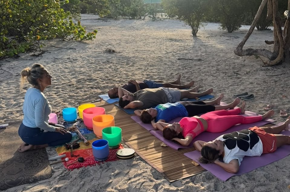 Uma mulher loira sentada na areia da praia, de perfil, com tigelas coloridas de cristal à frente, conduzindo uma meditação com seis pessoas deitadas em esteiras de yoga atrás dela