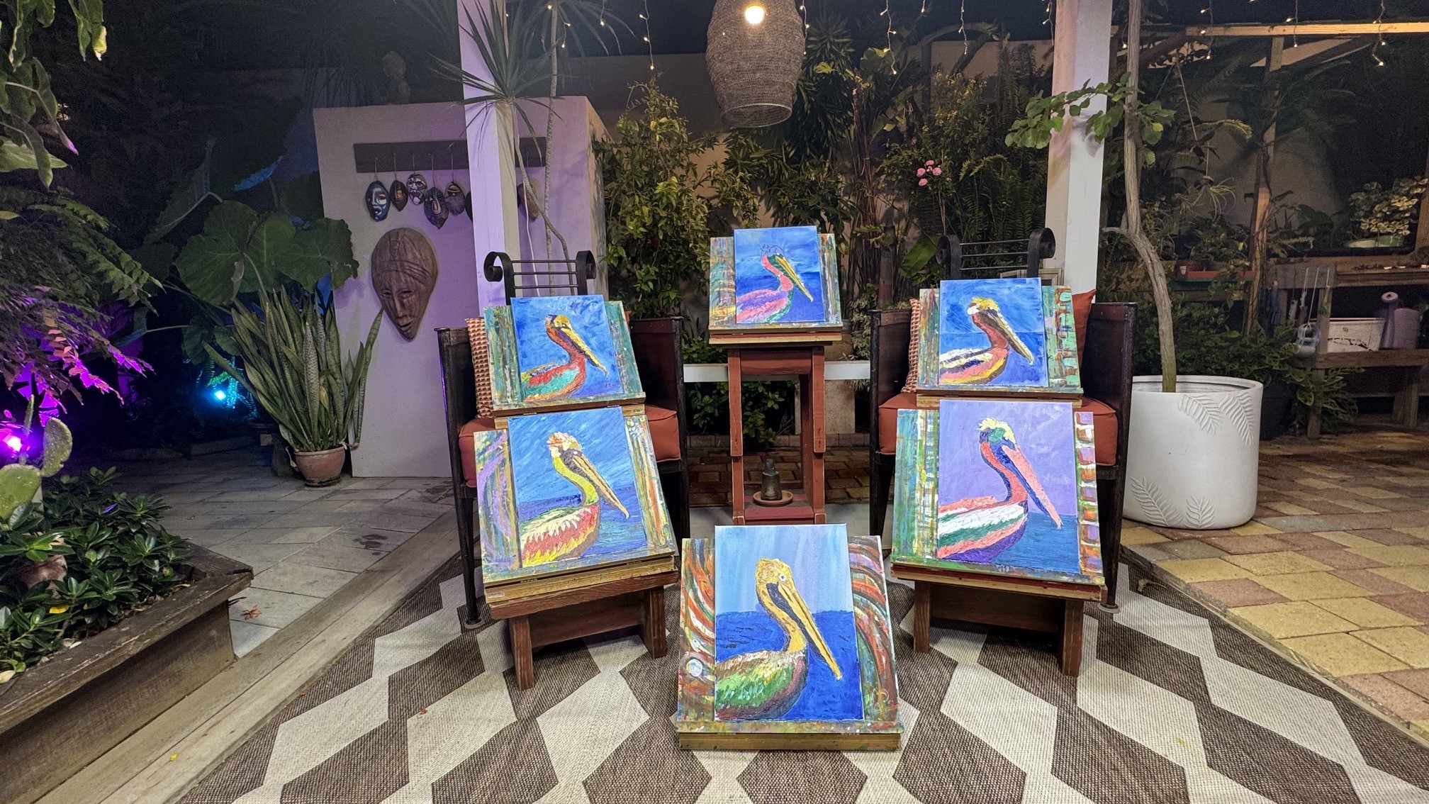 Oito pinturas de pelicanos coloridos em cavaletes de madeira, dispostas em um pátio externo com plantas e iluminação ambiente. Ao fundo, uma máscara tribal decora uma parede branca