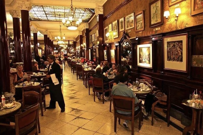 Interior de um caf&eacute; elegante com teto de vitral iluminado, colunas escuras e paredes com quadros. Clientes sentados em mesas redondas de madeira, enquanto um gar&ccedil;om de terno preto e gravata borboleta caminha pelo corredor central, segurando um pano branco